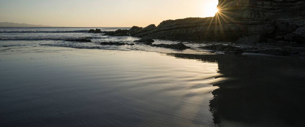 A beach with calm waters, a rocky cliff, and the sun setting on the horizon.