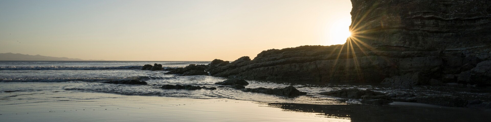 A beach with calm waters, a rocky cliff, and the sun setting on the horizon.