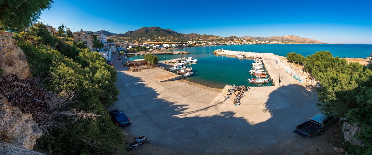 A coastal town with a marina, parked cars, and a mountainous backdrop.