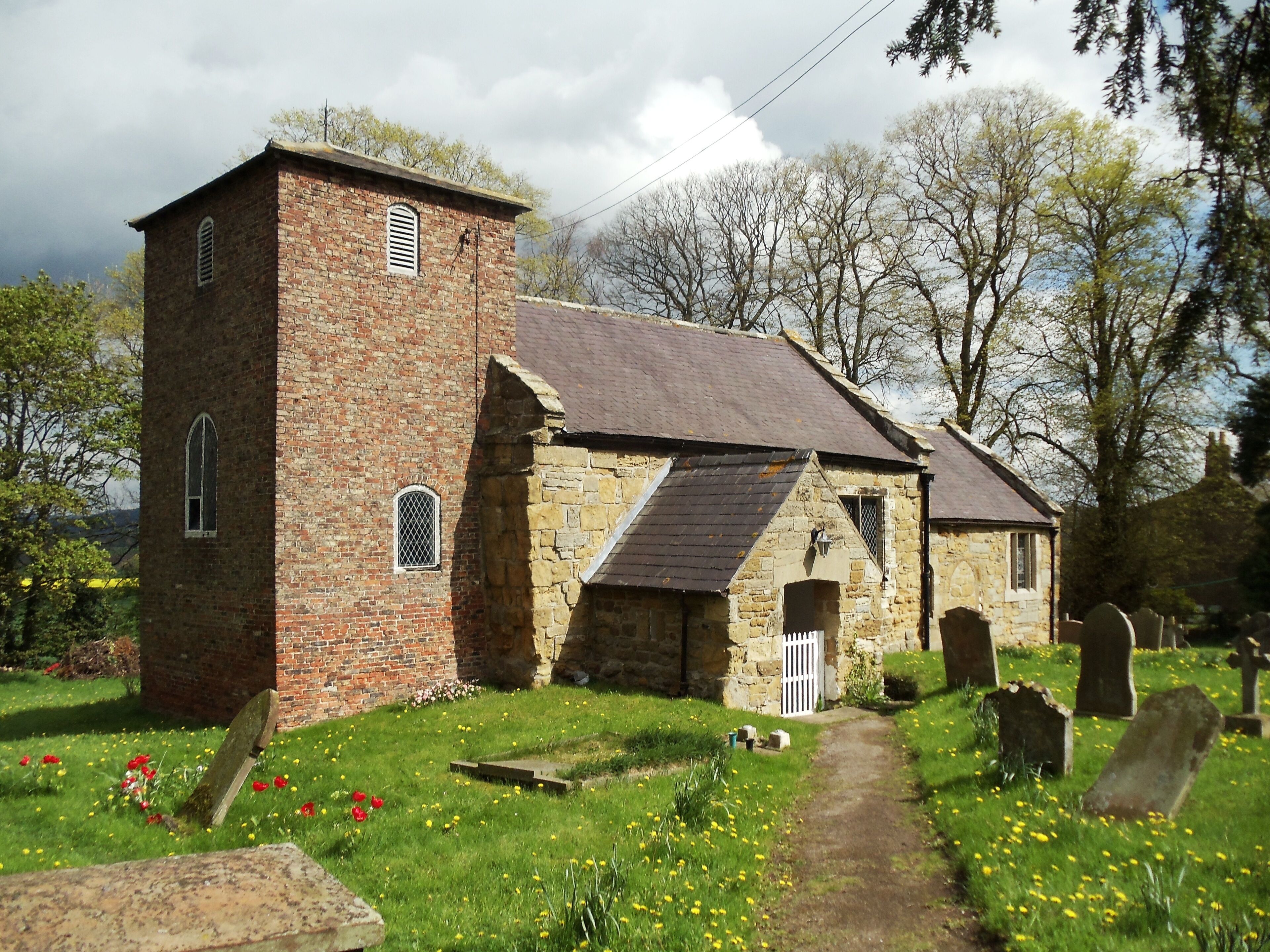 A church with a brick tower and stone walls, surrounded by a cemetery with headstones.