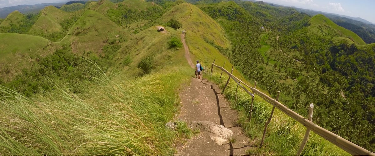 A hiking trail on a grassy hill with a wooden fence and a person walking.