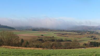 Rolling green fields, a mist-covered hill, and a clear blue sky.