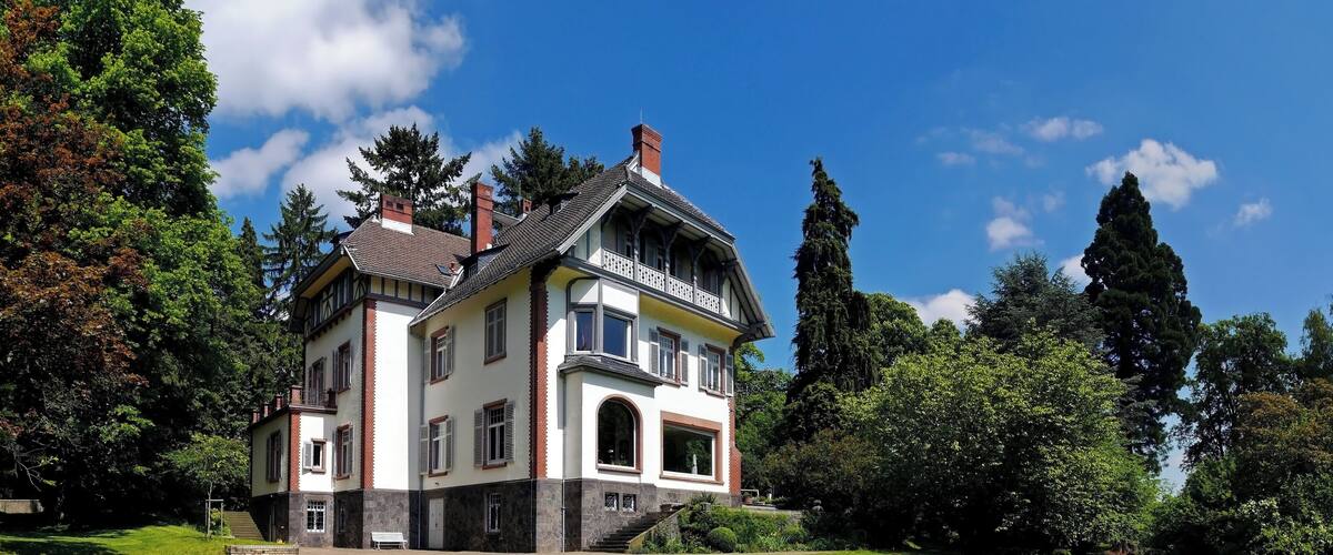 A large, two-story house with a white facade and red brick accents, surrounded by lush greenery and tall trees.