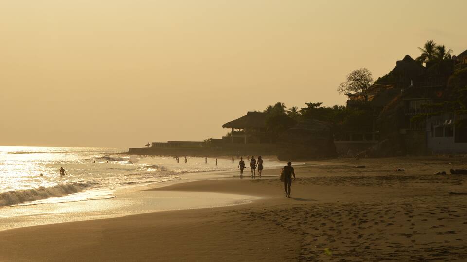 A beach with people walking along the shore, palm trees, and buildings in the background.