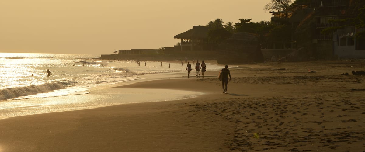 A beach with people walking along the shore, palm trees, and buildings in the background.