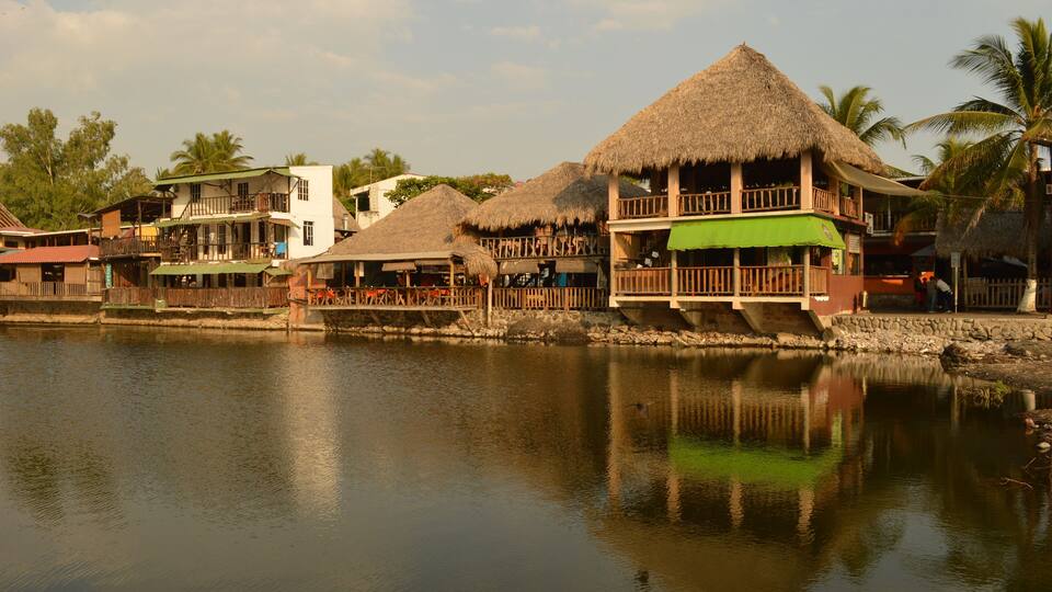 A waterfront with thatched-roof buildings and palm trees.