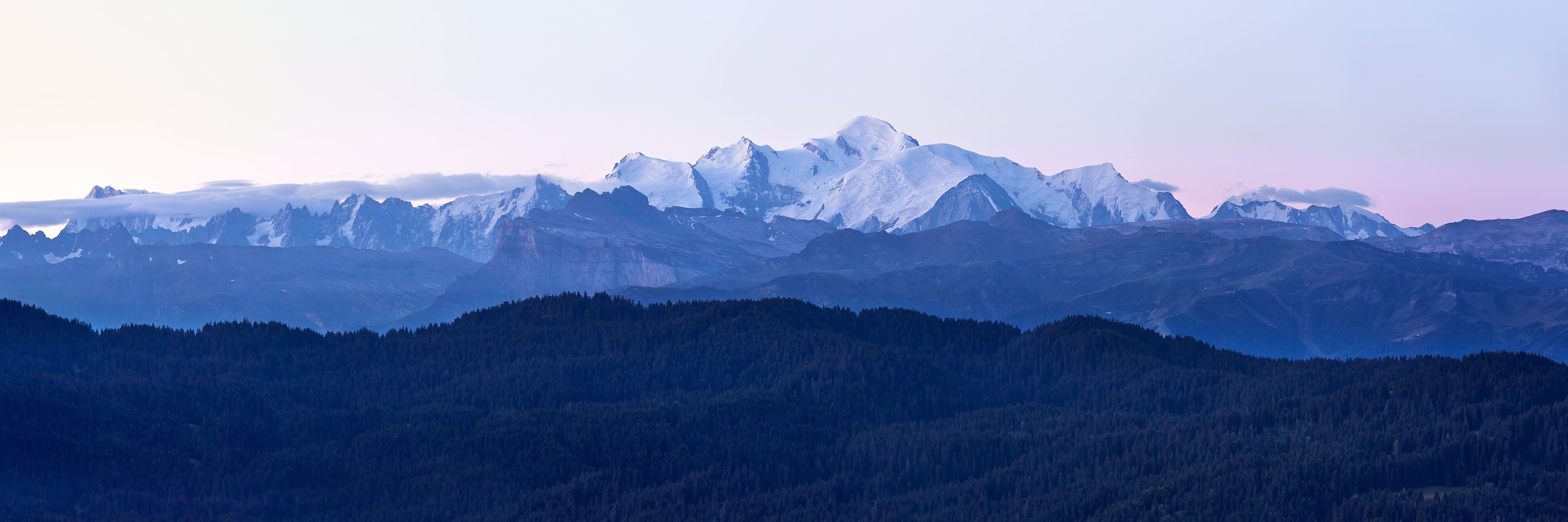 A range of snow-capped mountains with dense forests at their base.