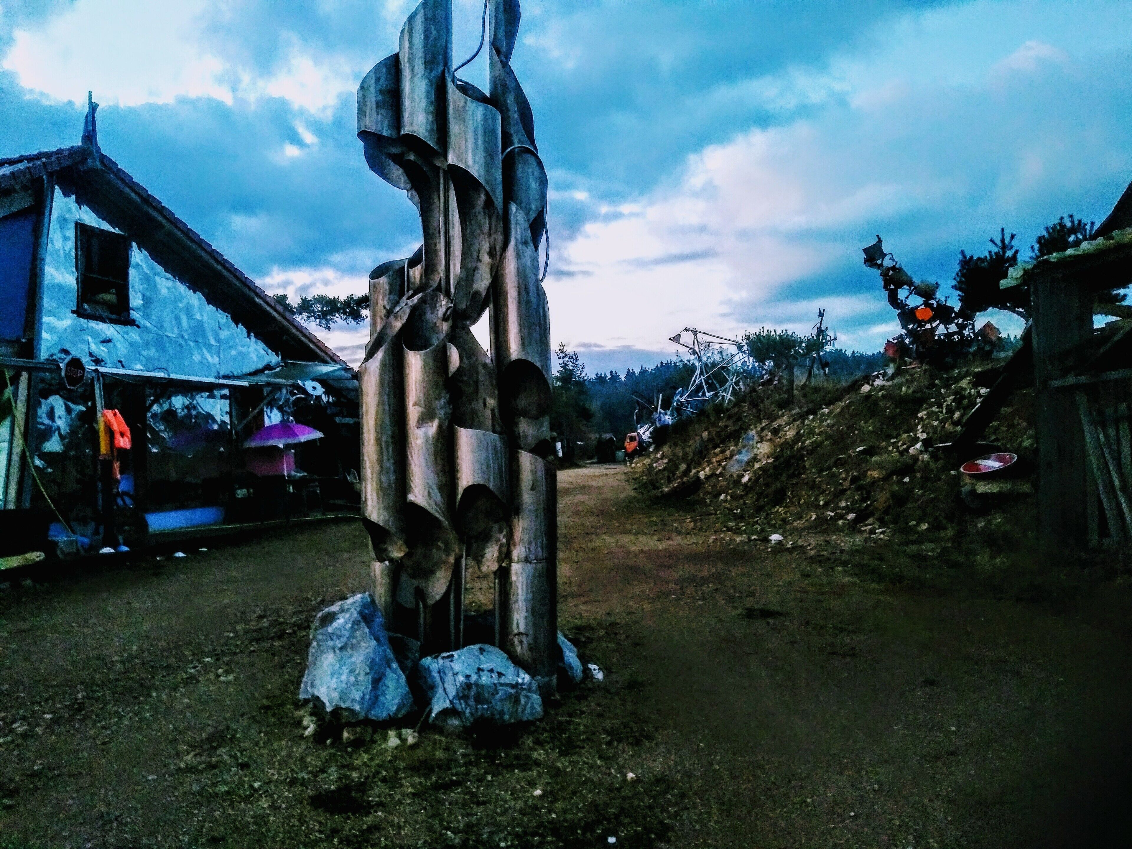 A rustic sculpture in a rural setting with a dirt road, houses, and a cloudy sky.