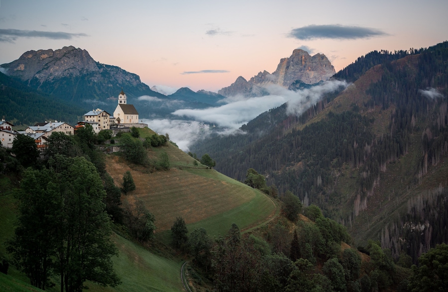Un paisaje montañoso con una iglesia en una colina, rodeado de bosques densos y un valle cubierto de niebla.