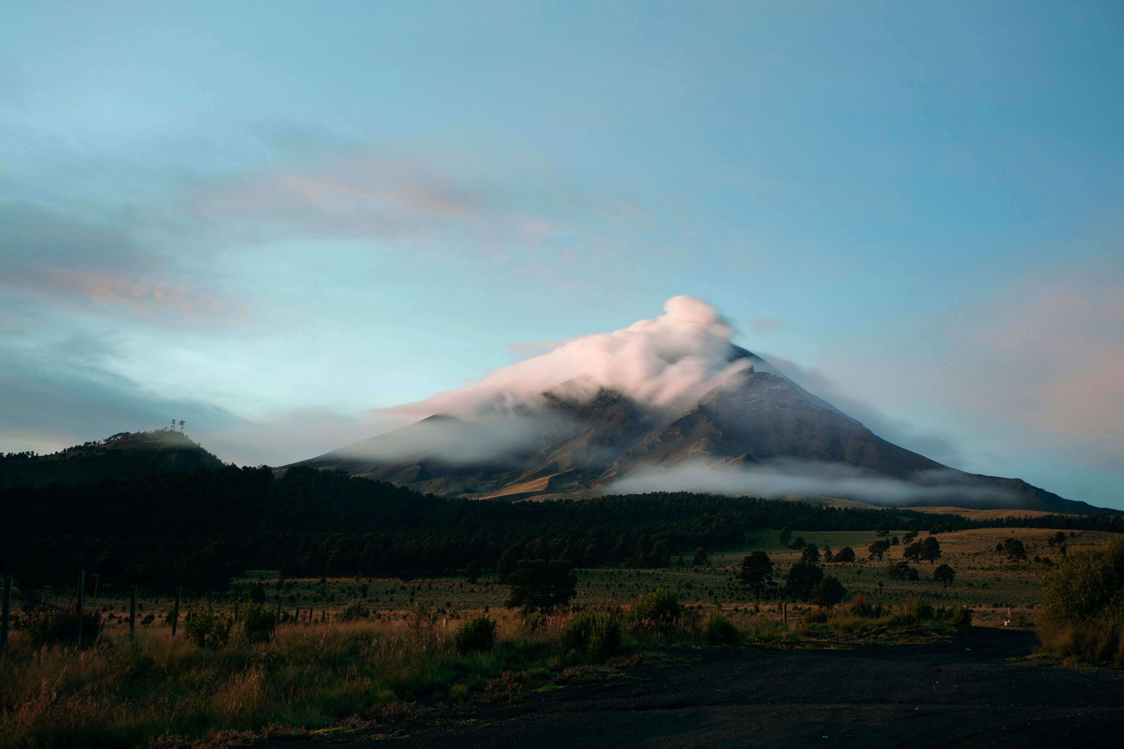 A mountain with a cloud base, surrounded by a grassy field and a fence.