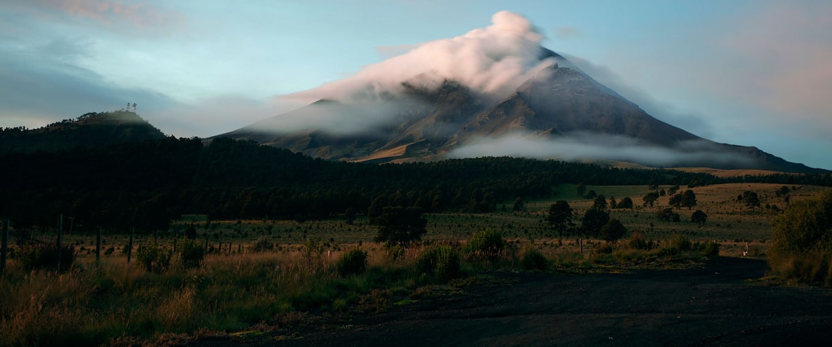 A mountain with a cloud base, surrounded by a grassy field and a fence.