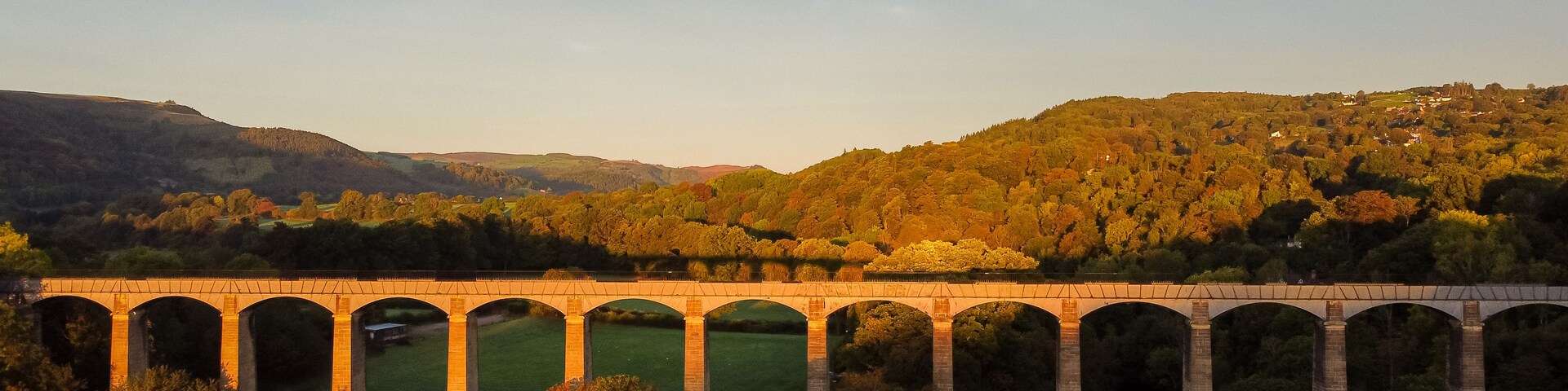Um viaduto de pedra sobre um rio, com uma encosta arborizada ao fundo.