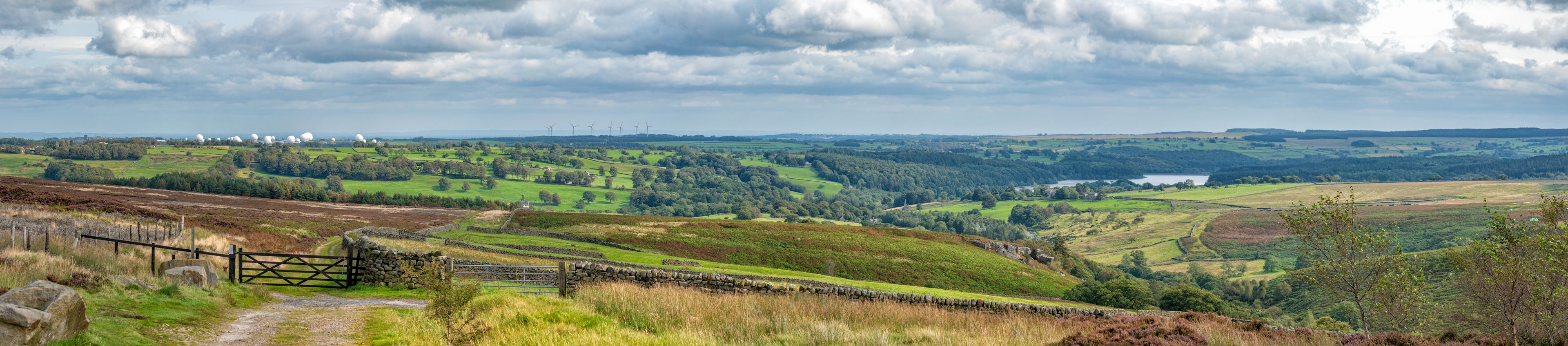 A rural landscape with rolling hills, a stone wall, a gate, and wind turbines in the distance.