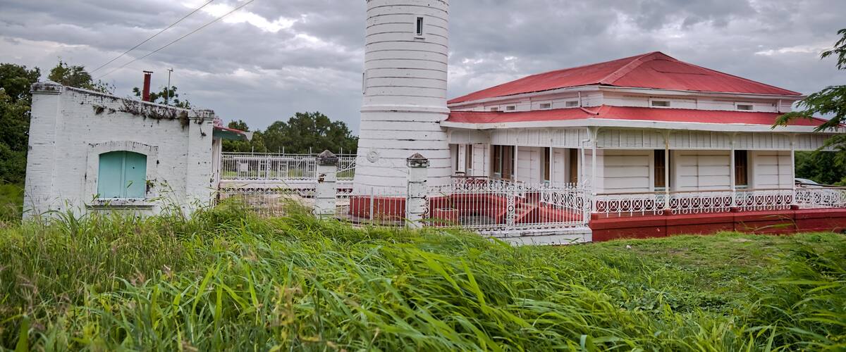 A white lighthouse with a red roof, surrounded by lush greenery and a cloudy sky.