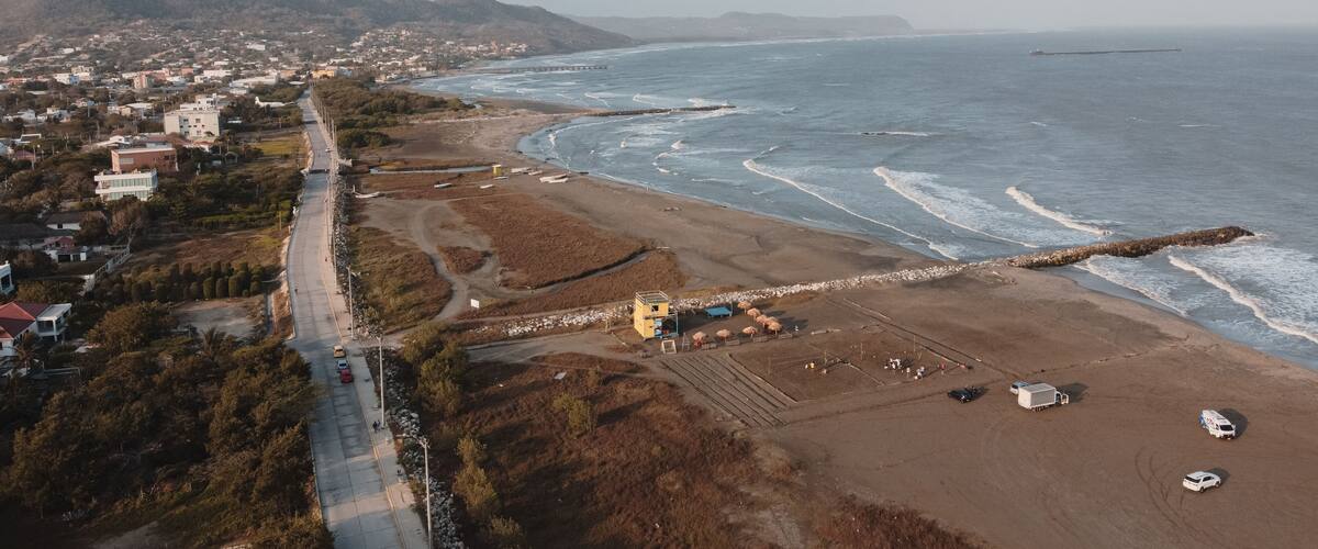 An aerial view of a coastal town with a beach, a road, and a distant mountain range.