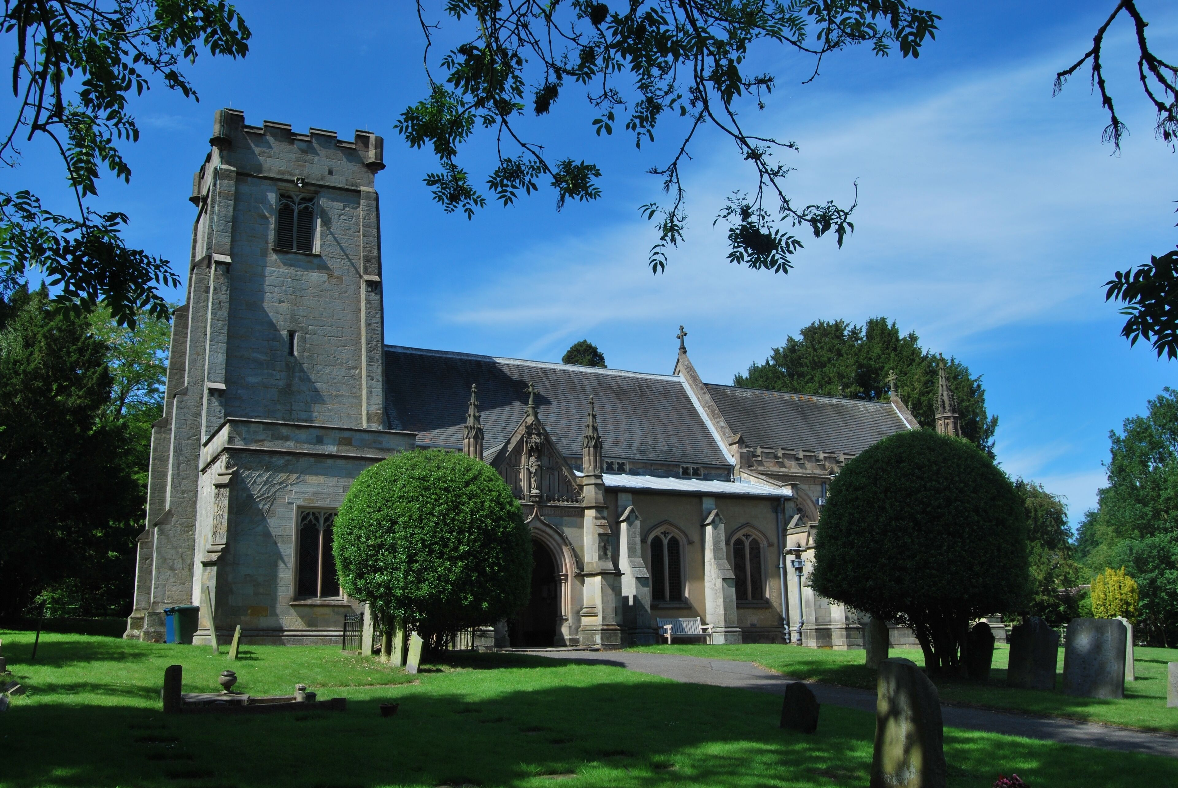 A historic church with a tall tower, surrounded by a cemetery with headstones and trimmed trees.