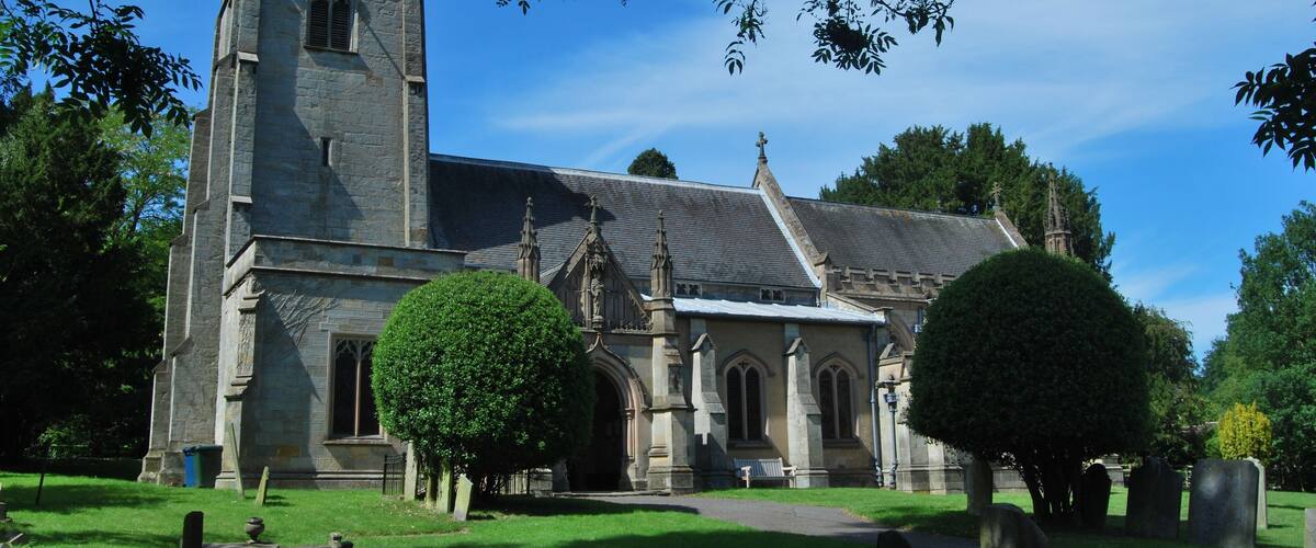 A historic church with a tall tower, surrounded by a cemetery with headstones and trimmed trees.