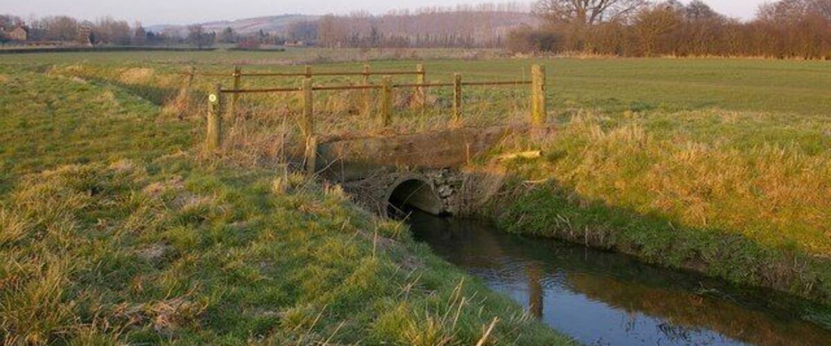 A rustic stone bridge over a narrow stream in a grassy field with distant trees and hills.