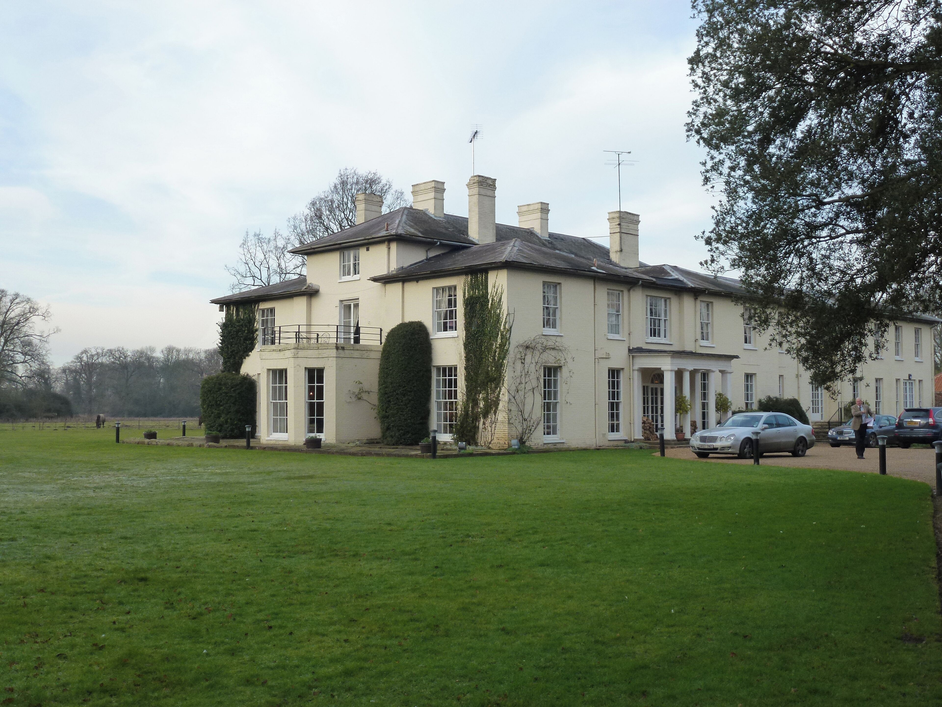 A large, cream-colored building with multiple chimneys and a balcony, surrounded by a well-maintained lawn and parked cars.