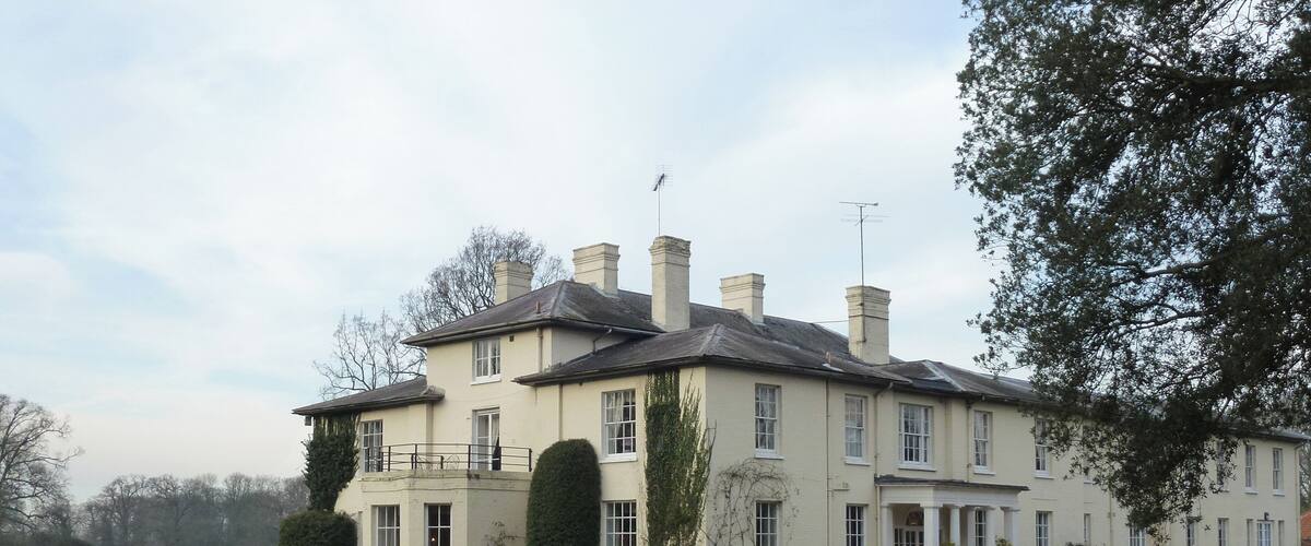 A large, cream-colored building with multiple chimneys and a balcony, surrounded by a well-maintained lawn and parked cars.
