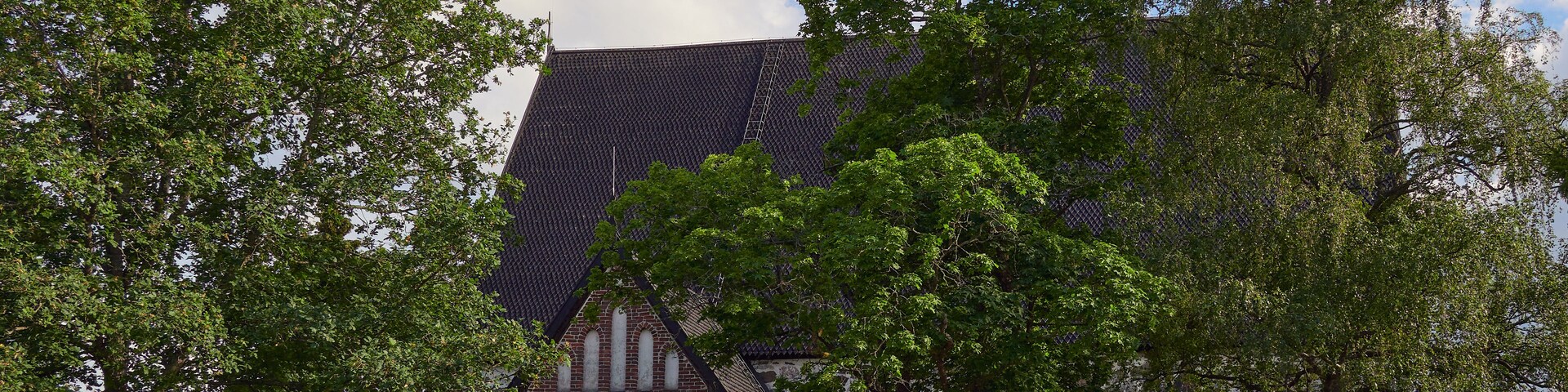 Um edifício histórico de pedra com telhado de azulejos, rodeado por árvores verdejantes e um muro de pedra.