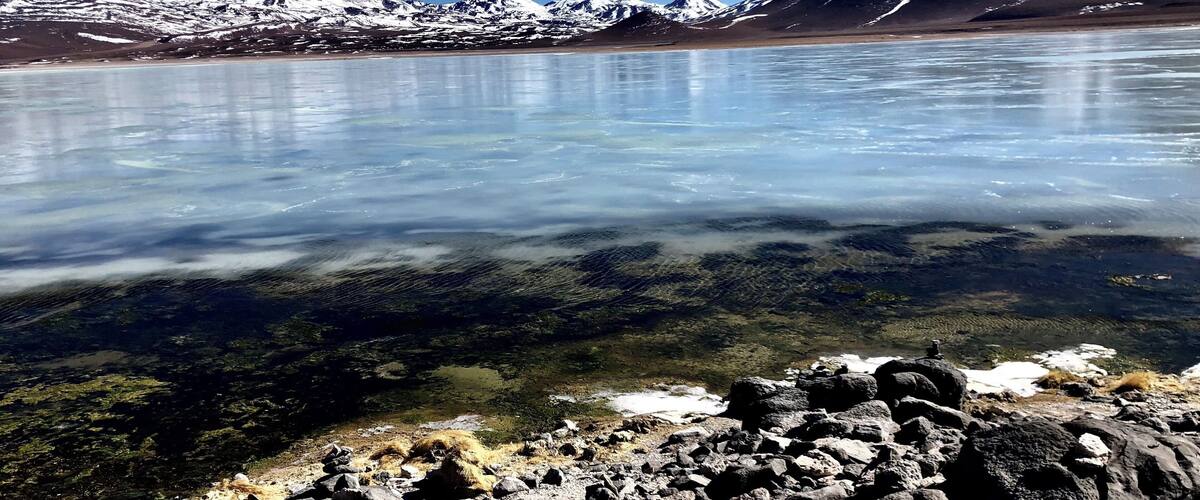 A frozen lake with clear water, surrounded by rocky terrain and snow-capped mountains.