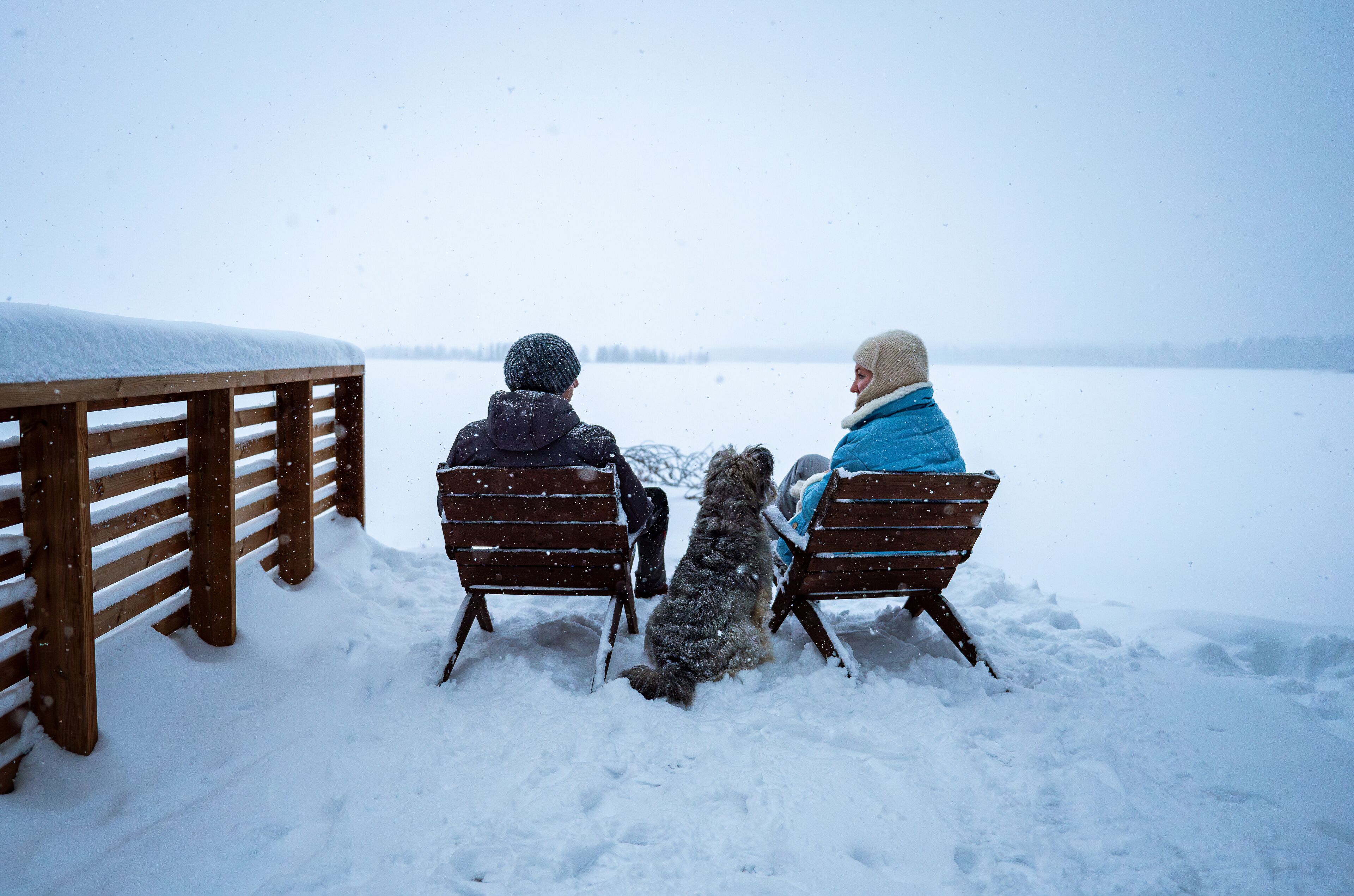 Couple with dog sitting on snowy lakeside enjoying winter scenery.