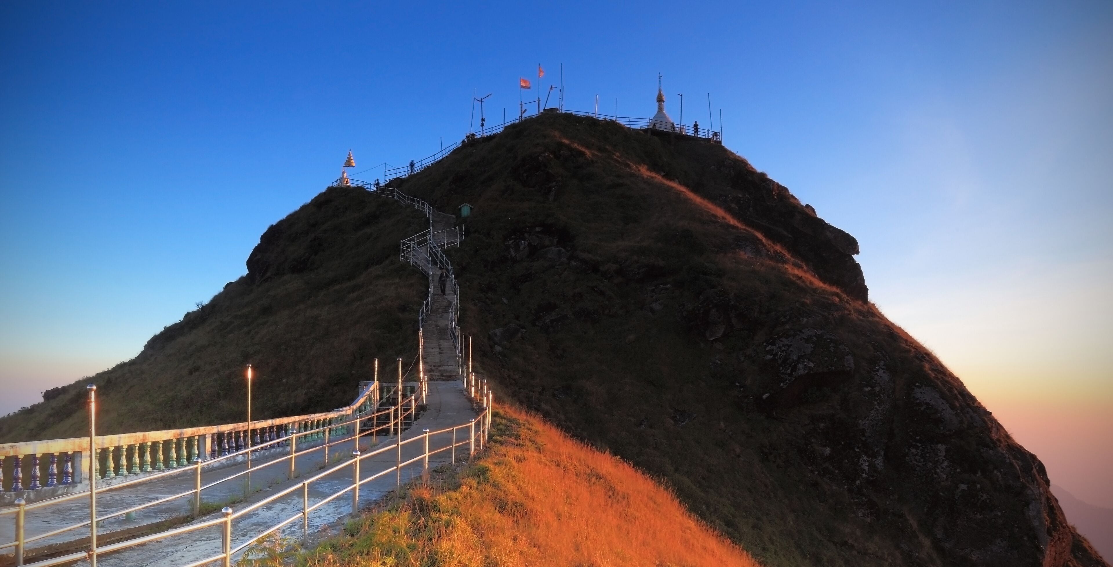 A mountain with a shrine at the peak, a staircase leading up, and a guardrail along a path.