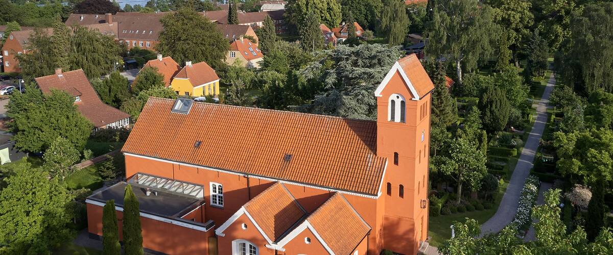 A red brick church with a bell tower, surrounded by lush greenery and a clear view of a river in the background.
