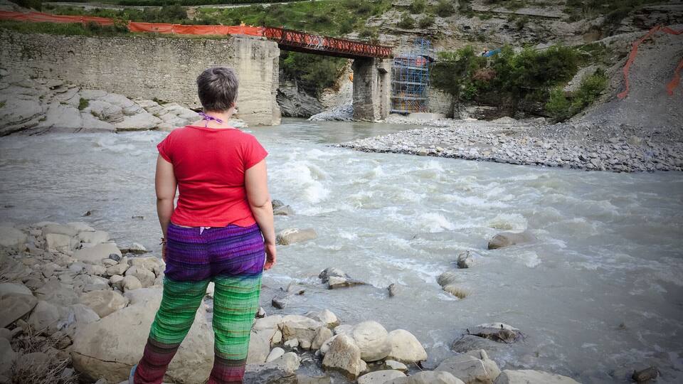 A person standing by a river with a bridge in the background.