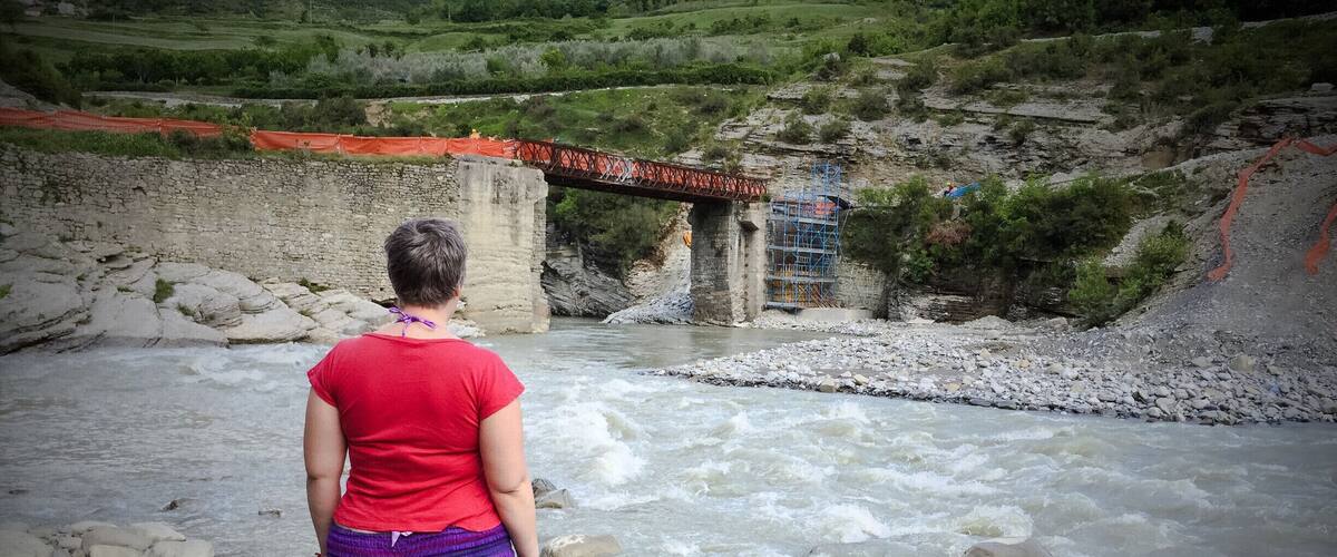 A person standing by a river with a bridge in the background.