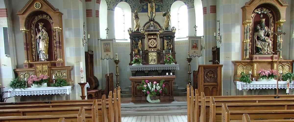 An aisle with wooden pews and a patterned floor leading to an altar with statues and floral arrangements.