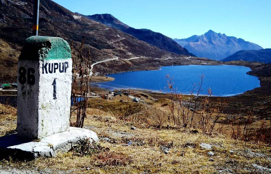 A mountain landscape with a lake, a signpost with '86 KUPUP 1', and distant mountains.