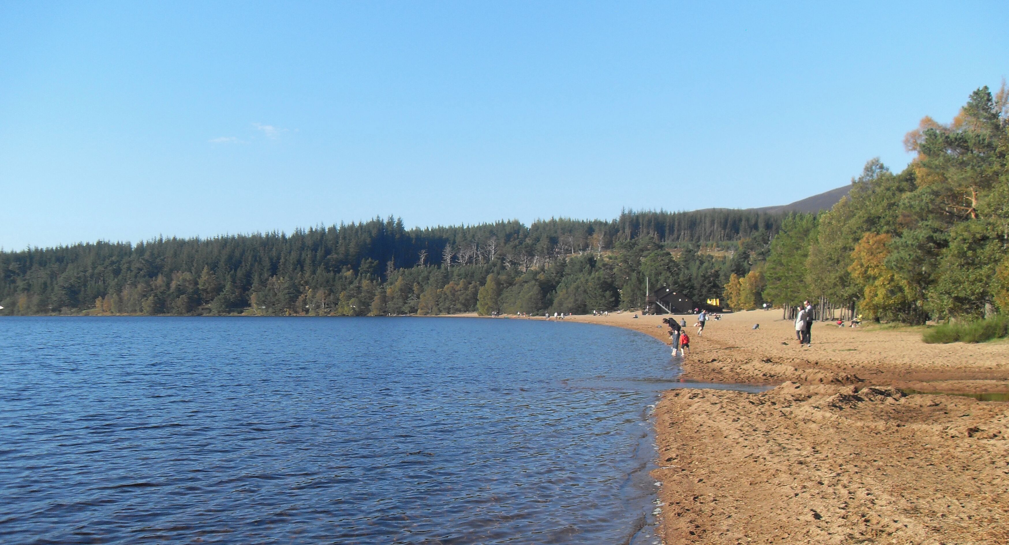 A lake with a sandy beach, people walking, and a forested hill in the background.