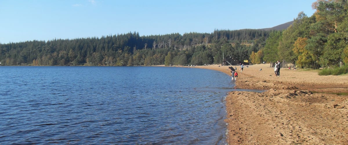 A lake with a sandy beach, people walking, and a forested hill in the background.