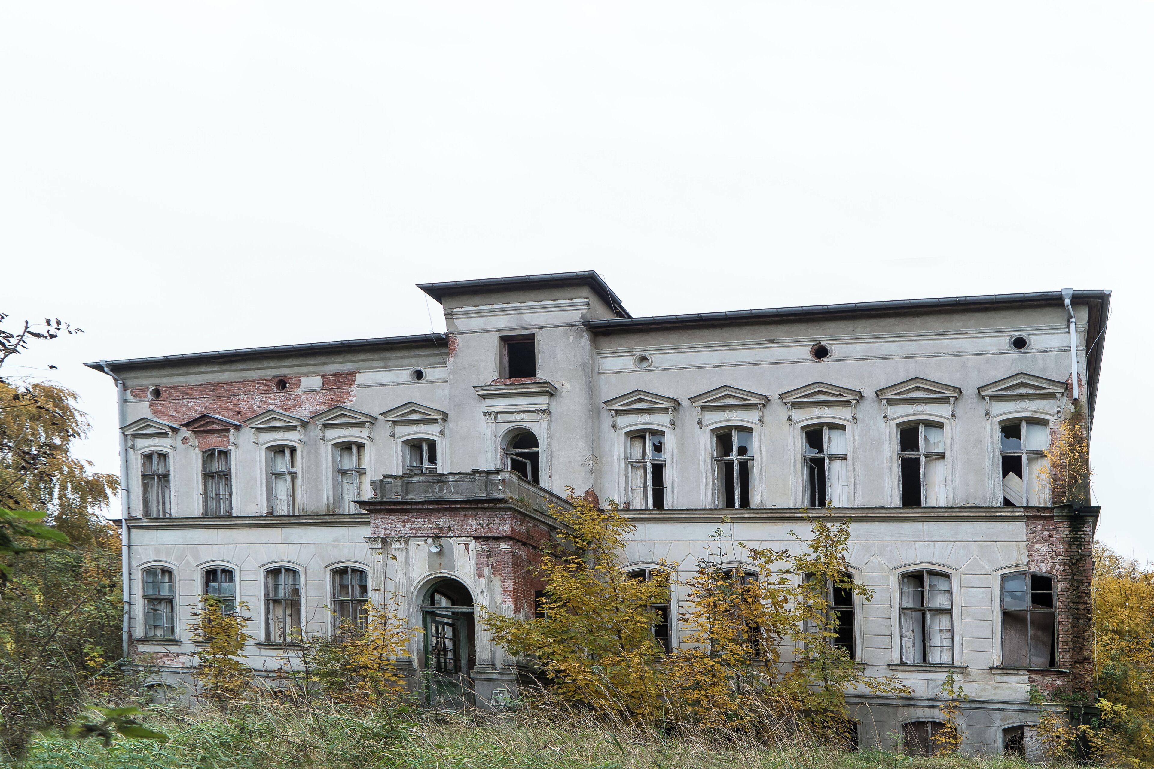 A large, historic building with multiple windows and a balcony, surrounded by overgrown vegetation.