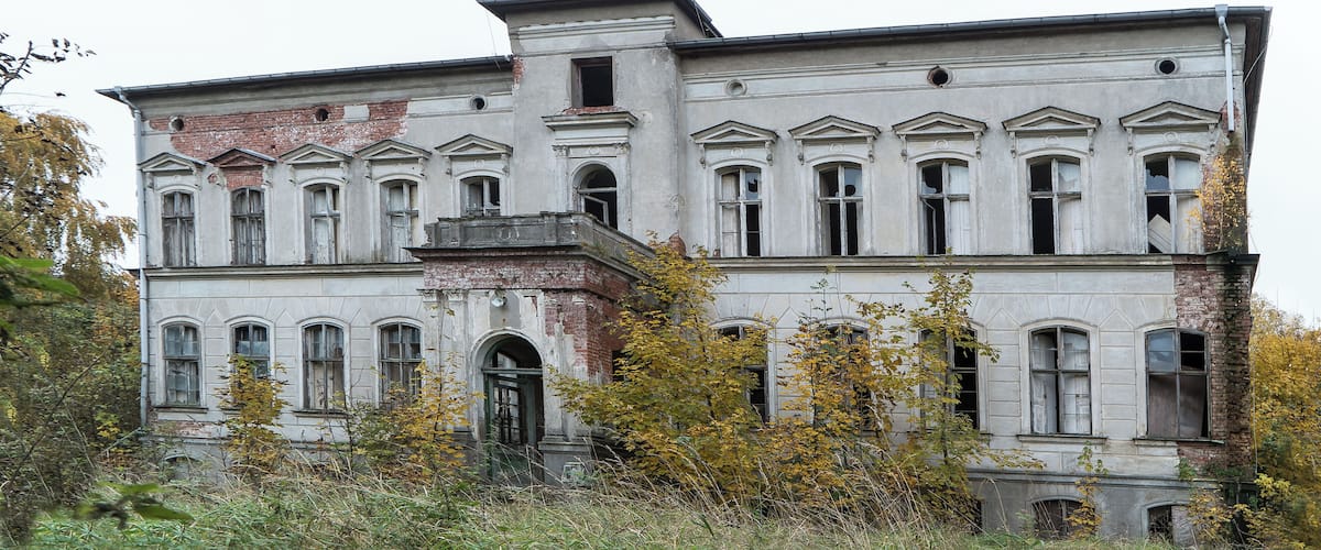 A large, historic building with multiple windows and a balcony, surrounded by overgrown vegetation.