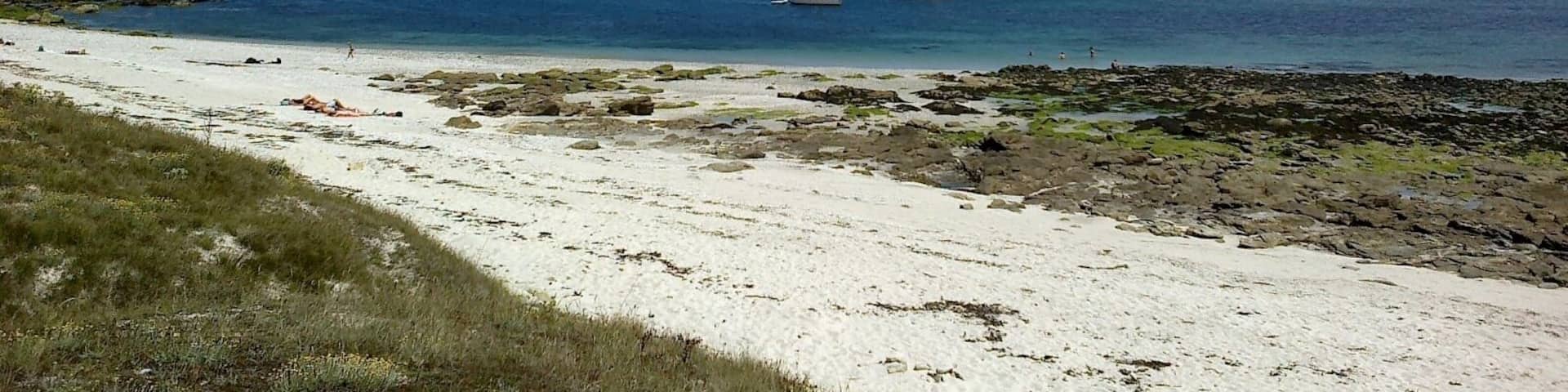 A beach with white sand and rocky areas, and several sailboats anchored in the water.