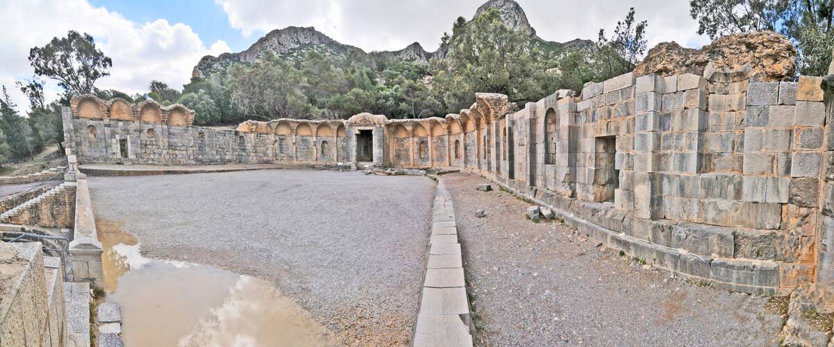An ancient stone structure with arched doorways and a large open area, surrounded by a stone wall and a mountainous backdrop.