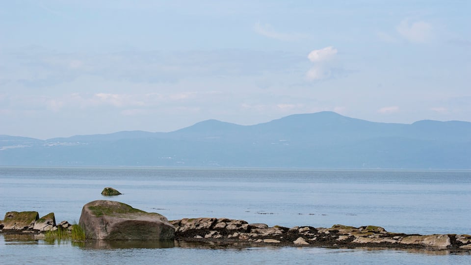 A calm lake with a rocky shoreline and distant mountains under a clear sky.
