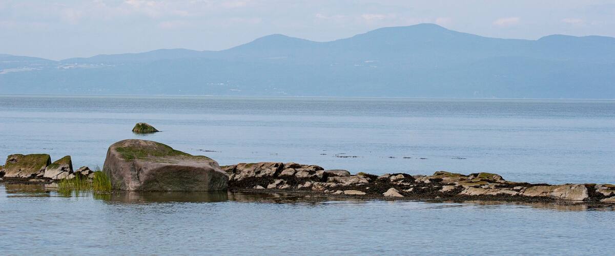 A calm lake with a rocky shoreline and distant mountains under a clear sky.