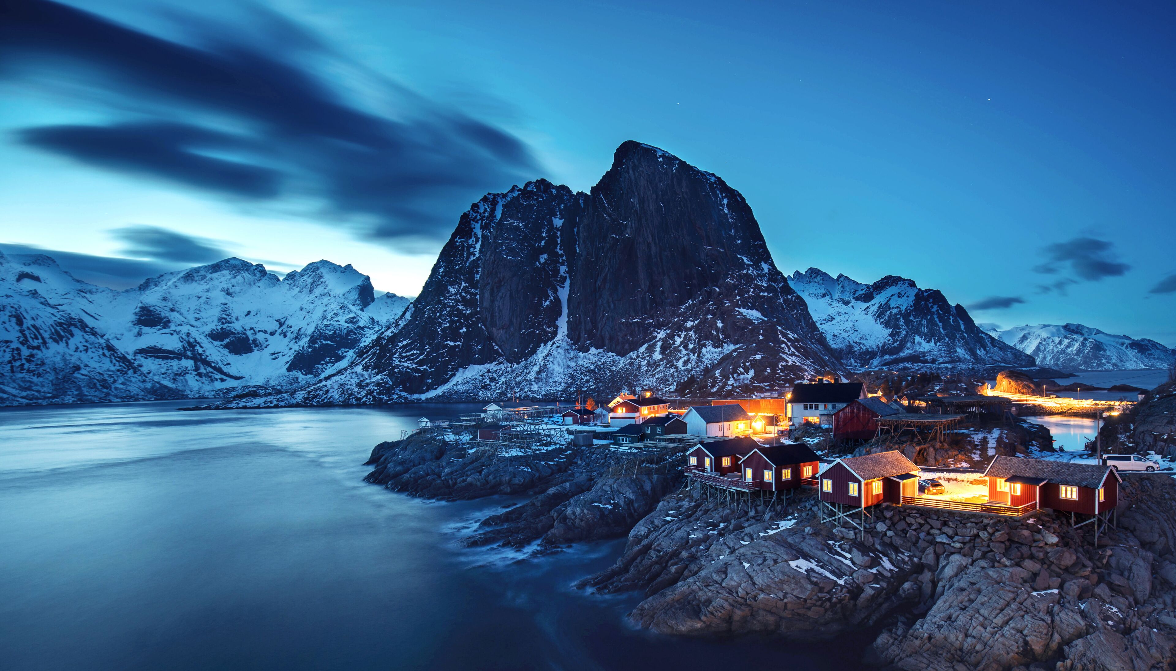 A fjord with a prominent mountain, snow-covered peaks, and a small settlement with illuminated buildings.