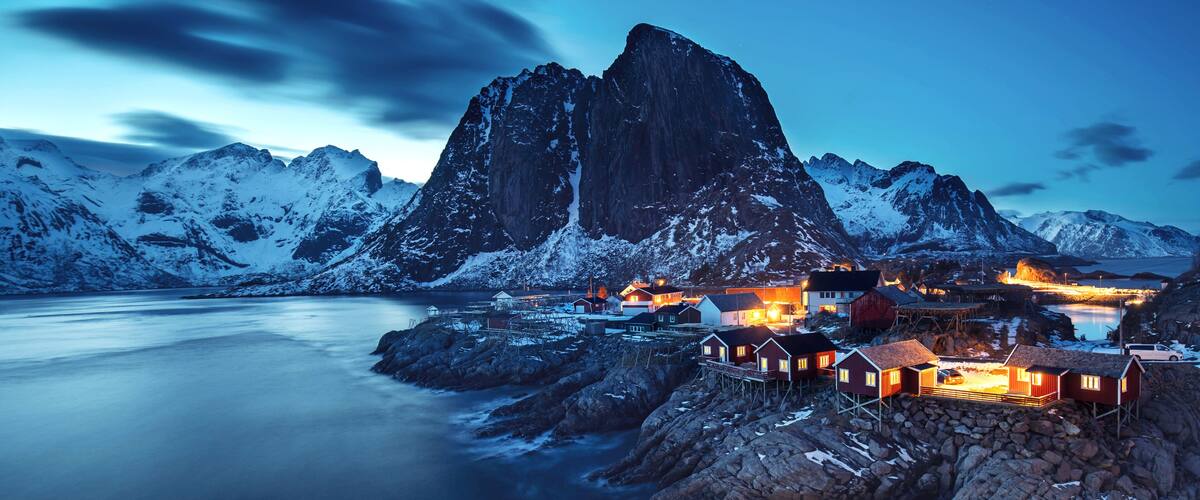 A fjord with a prominent mountain, snow-covered peaks, and a small settlement with illuminated buildings.
