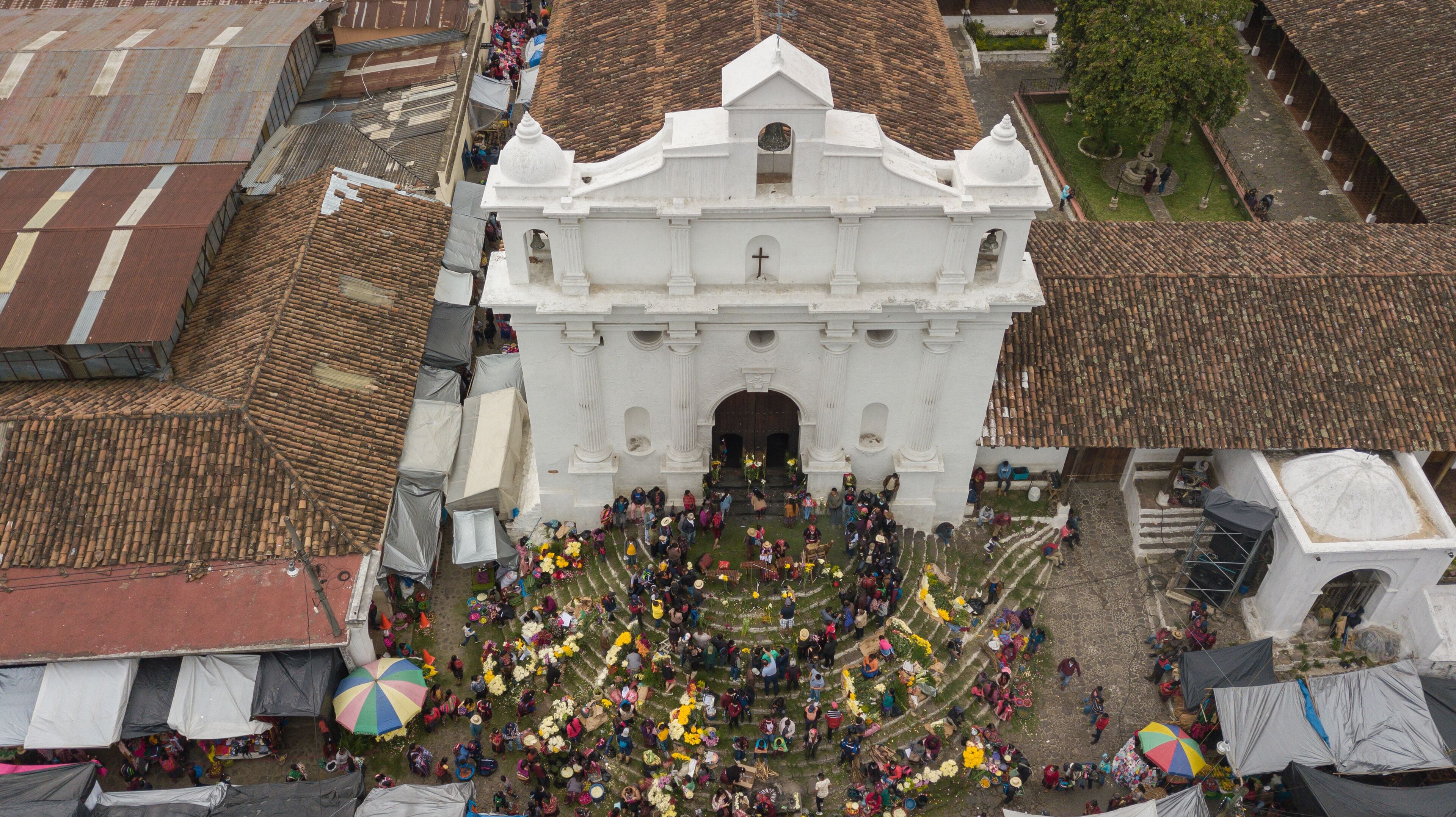 An aerial view of a large white church with a tiled roof, surrounded by a crowd of people and colorful umbrellas.