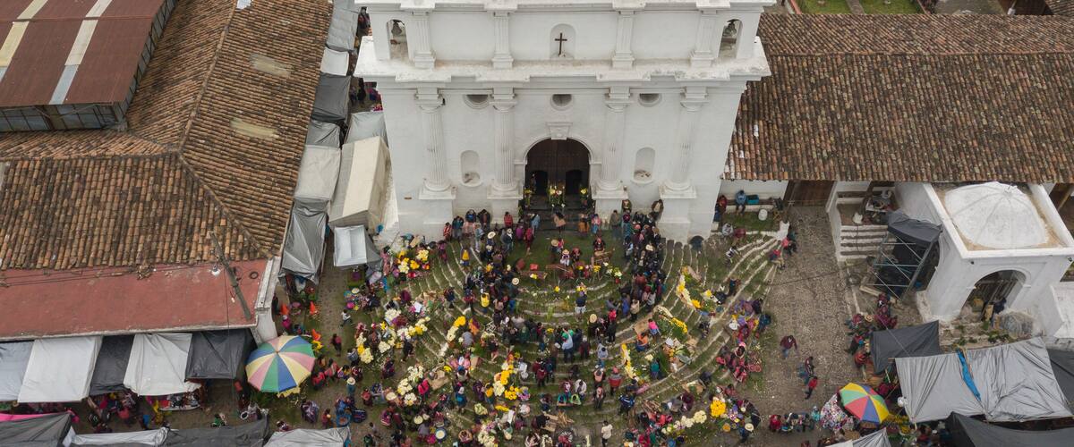 An aerial view of a large white church with a tiled roof, surrounded by a crowd of people and colorful umbrellas.