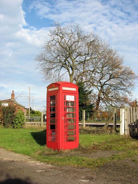 Uma cabine telefônica vermelha em área rural, com uma árvore e uma cerca ao fundo.