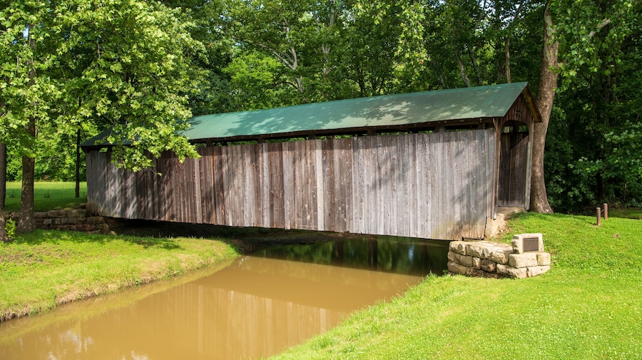 A wooden covered bridge over a calm river, surrounded by lush greenery and trees.