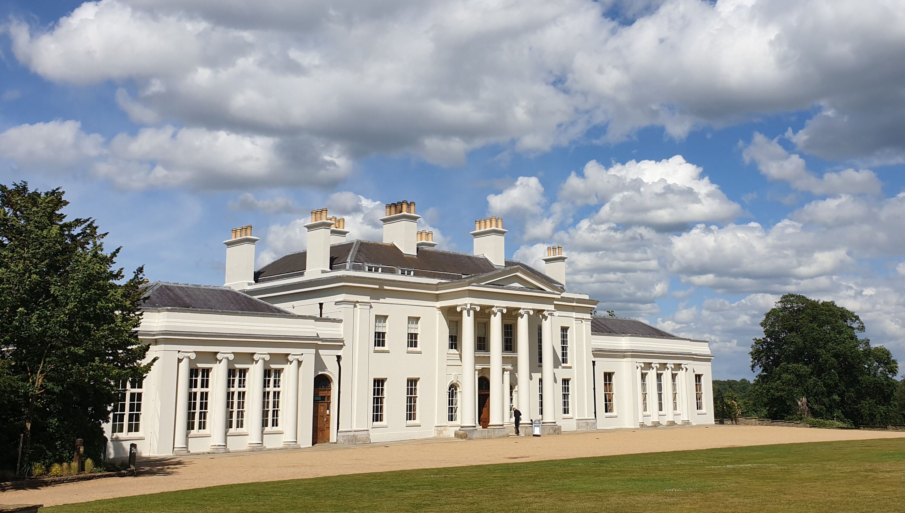 A large white mansion with multiple chimneys and columns, surrounded by a well-maintained lawn and trees.