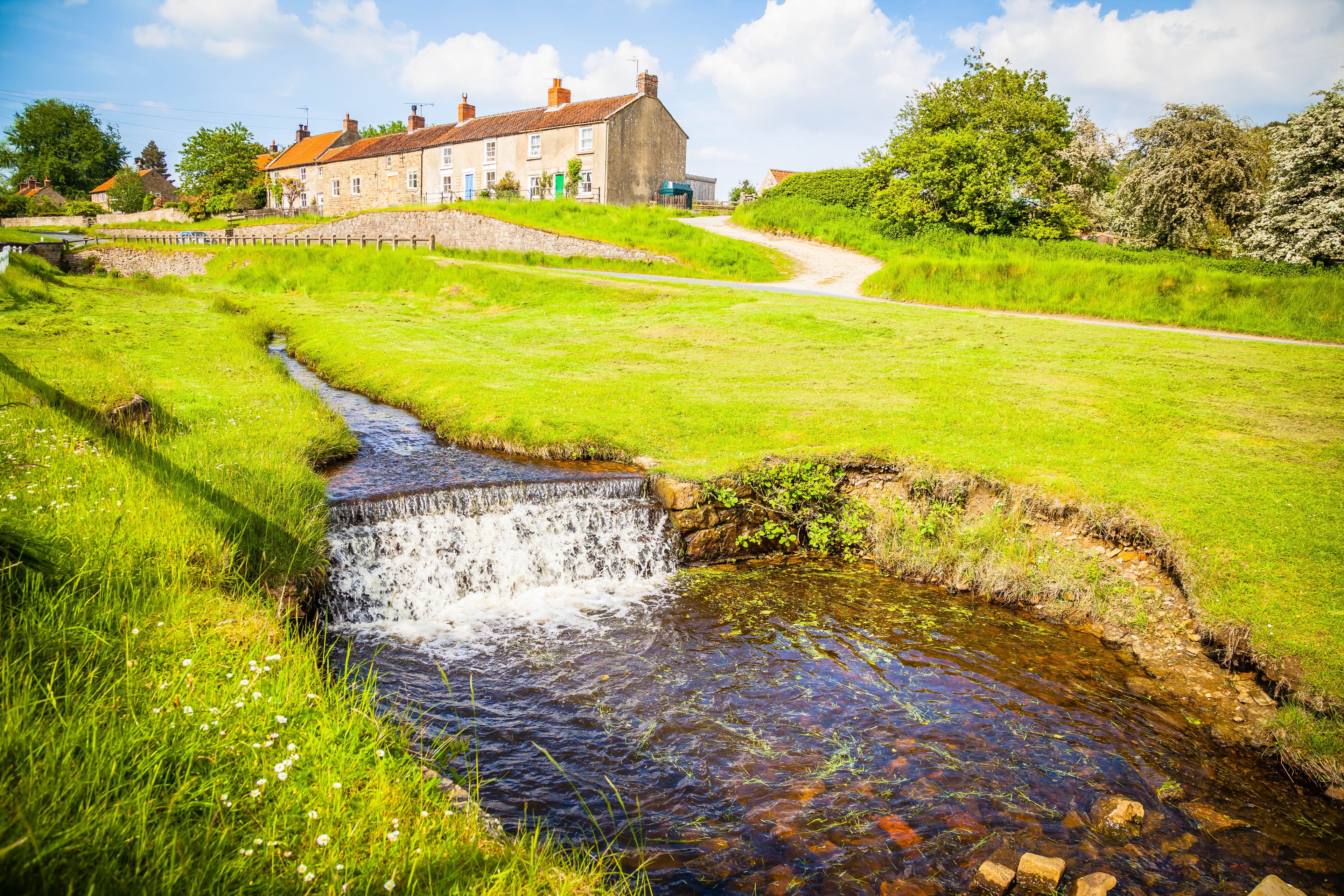 A rural landscape with a small stream, green fields, and a stone building in the background.