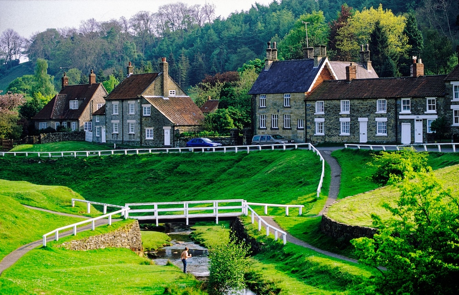 Un paisaje verde exuberante con una cerca blanca, edificios de piedra y un río.